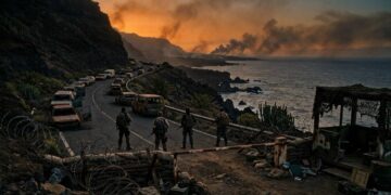 Retén militar en una carretera costera volcánica al atardecer, con autos abandonados y humo en el horizonte