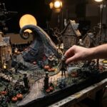 Maqueta de stop motion de un pueblo gótico con calabazas y una colina en espiral, con la mano de un animador moviendo un muñeco
