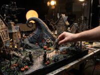 Maqueta de stop motion de un pueblo gótico con calabazas y una colina en espiral, con la mano de un animador moviendo un muñeco
