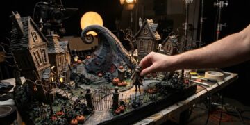 Maqueta de stop motion de un pueblo gótico con calabazas y una colina en espiral, con la mano de un animador moviendo un muñeco