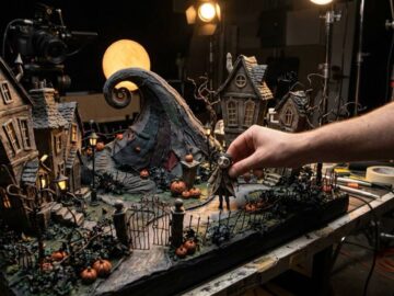 Maqueta de stop motion de un pueblo gótico con calabazas y una colina en espiral, con la mano de un animador moviendo un muñeco