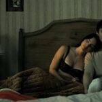Una pareja en una habitación con papel tapiz antiguo y un reloj despertador, en una escena de terror romántico